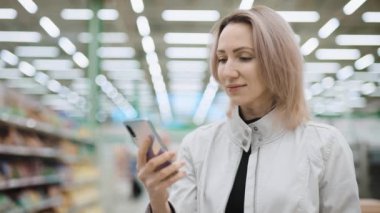 Süpermarketteki güzel kadın akıllı telefon kullanıyor. Bir müşteri mobil uygulama kullanarak fiyatları karşılaştırır. Alışveriş teknolojileri.