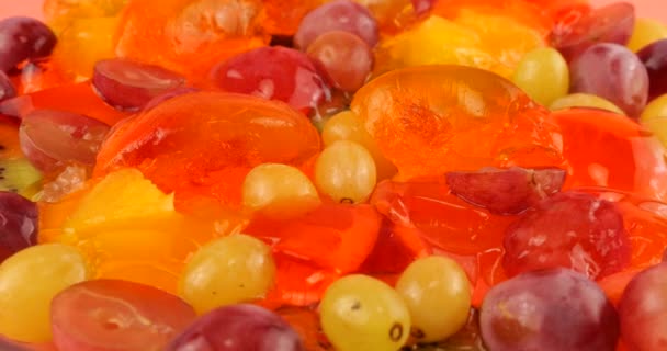 マルチカラーのベリーゼラチンゼリーにオレンジとキウイのスライスをピンクの背景に落ちるブドウ — ストック動画
