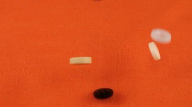 Plastic multi-colored buttons fall on an orange cotton fabric. Slow motion.