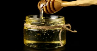 Honey flows down from a spoon spindle into a jar with honey on a black background.