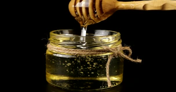 蜂蜜はスプーンのスピンドルから黒い背景の蜂蜜と瓶に流れ落ちる — ストック動画