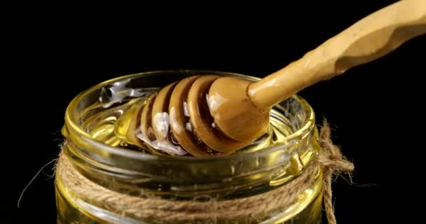 蜂蜜はスプーンのスピンドルから黒い背景の蜂蜜と瓶に流れ落ちる — ストック動画