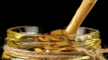Honey flows down from a spoon spindle into a jar with honey on a black background.