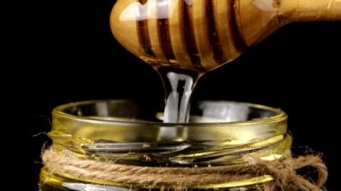 Honey flows down from a spoon spindle into a jar with honey on a black background.