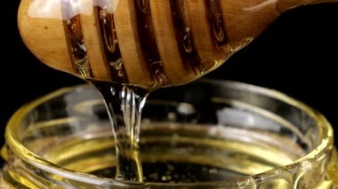 Honey flows down from a spoon spindle into a jar with honey on a black background.