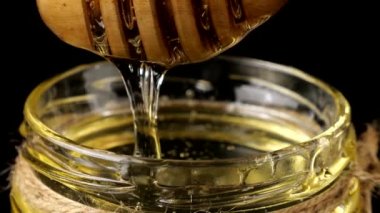 Honey flows down from a spoon spindle into a jar with honey on a black background.