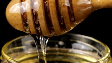 Honey flows down from a spoon spindle into a jar with honey on a black background.