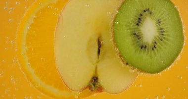 Slices of ripe orange, apple and kiwi in transparent water in air bubbles on an orange background.