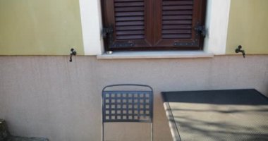 Bahçe metali mobilyalar, masa ve sandalyeler, ev duvarı ve panjurlu pencereler.