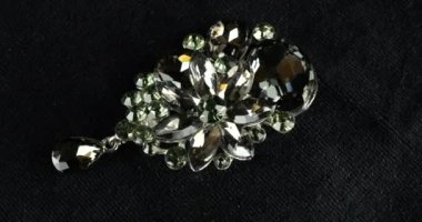 Çiçek buketi şeklinde gümüş renkli klasik broş, kadife vitrin üzerinde gri ve yeşil kristaller var..