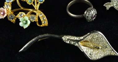Eski mücevherlerle birlikte kadife kaplama vitrin: Broşlar ve yüzükler. Altın ve gümüş renkli mücevherler, çok renkli kristaller ve metal tanecikleri var..