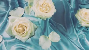 Beyaz güllerin çiçekleri ve taç yaprakları mavi ipek arka planda suyun yüzeyinde yüzerler..