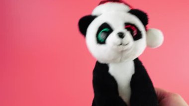 Doldurulmuş ve pofuduk oyuncak panda Noel Baba şapkasıyla pembe arka planda dans ediyor..