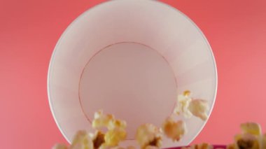 Patlamış mısır taneleri pembe arka planda izole edilmiş kırmızı ve beyaz çizgili kovaya düşer. Atıştırmalık film, fast food kafe tatlı aperatif. Üst Manzara. Yavaş çekim.