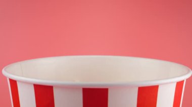 Patlamış mısır taneleri pembe arka planda izole edilmiş kırmızı ve beyaz çizgili kovaya düşer. Atıştırmalık film, fast food kafe tatlı aperatif. Yavaş çekim.