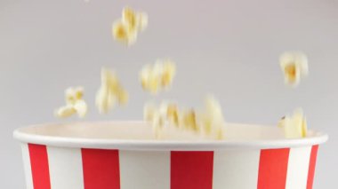 Patlamış mısır taneleri beyaz arka planda izole edilmiş kırmızı ve beyaz çizgili kovaya düşüyor. Atıştırmalık film, fast food kafe tatlı aperatif. Yavaş çekim.