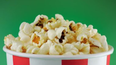 Patlamış mısır taneleri yeşil arka planda izole edilmiş kırmızı ve beyaz çizgili kovada dönüyorlar. Atıştırmalık film, fast food kafe tatlı aperatif. Kromakey veya krom anahtar arkaplanı.