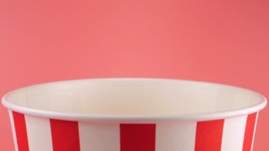 Patlamış mısır taneleri pembe arka planda izole edilmiş kırmızı ve beyaz çizgili kovaya düşer. Atıştırmalık film, fast food kafe tatlı aperatif. Yavaş çekim.