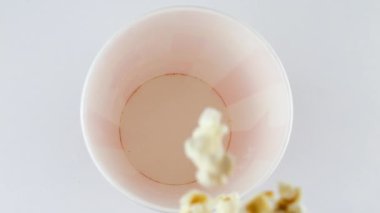 Patlamış mısır taneleri beyaz arka planda izole edilmiş kırmızı ve beyaz çizgili kovaya düşüyor. Atıştırmalık film, fast food kafe tatlı aperatif. Ağır çekim. Üst görünüm.