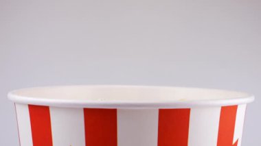 Patlamış mısır taneleri beyaz arka planda izole edilmiş kırmızı ve beyaz çizgili kovaya düşüyor. Atıştırmalık film, fast food kafe tatlı aperatif. Yavaş çekim.