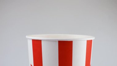 Patlamış mısır taneleri beyaz arka planda izole edilmiş kırmızı ve beyaz çizgili kovaya düşüyor. Atıştırmalık film, fast food kafe tatlı aperatif. Yavaş çekim.