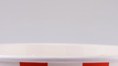 Patlamış mısır taneleri beyaz arka planda izole edilmiş kırmızı ve beyaz çizgili kovaya düşüyor. Atıştırmalık film, fast food kafe tatlı aperatif. Yavaş çekim.