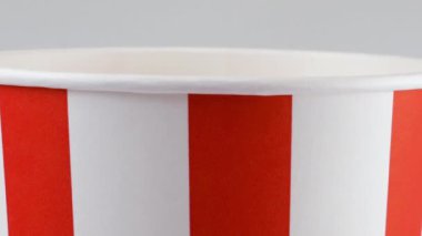Patlamış mısır taneleri beyaz arka planda izole edilmiş kırmızı ve beyaz çizgili kovaya düşüyor. Atıştırmalık film, fast food kafe tatlı aperatif. Yavaş çekim.