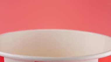 Patlamış mısır taneleri pembe arka planda izole edilmiş kırmızı ve beyaz çizgili kovaya düşer. Atıştırmalık film, fast food kafe tatlı aperatif. Yavaş çekim.