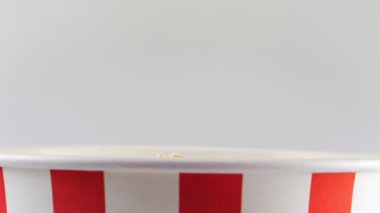 Patlamış mısır taneleri beyaz arka planda izole edilmiş kırmızı ve beyaz çizgili kovaya düşüyor. Atıştırmalık film, fast food kafe tatlı aperatif. Yavaş çekim.