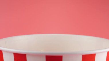Patlamış mısır taneleri pembe arka planda izole edilmiş kırmızı ve beyaz çizgili kovaya düşer. Atıştırmalık film, fast food kafe tatlı aperatif. Yavaş çekim.