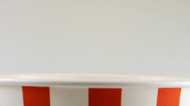 Patlamış mısır taneleri beyaz arka planda izole edilmiş kırmızı ve beyaz çizgili kovaya düşüyor. Atıştırmalık film, fast food kafe tatlı aperatif. Yavaş çekim.