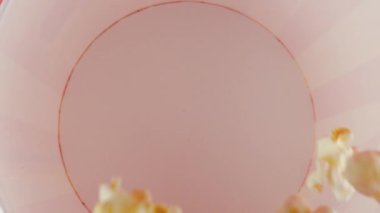 Patlamış mısır taneleri pembe arka planda izole edilmiş kırmızı ve beyaz çizgili kovaya düşer. Atıştırmalık film, fast food kafe tatlı aperatif. Üst Manzara. Yavaş çekim.