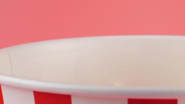 Patlamış mısır taneleri pembe arka planda izole edilmiş kırmızı ve beyaz çizgili kovaya düşer. Atıştırmalık film, fast food kafe tatlı aperatif. Yavaş çekim.