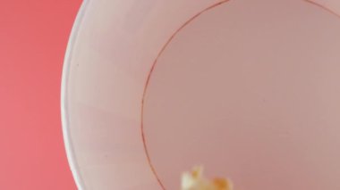Patlamış mısır taneleri pembe arka planda izole edilmiş kırmızı ve beyaz çizgili kovaya düşer. Atıştırmalık film, fast food kafe tatlı aperatif. Üst Manzara. Yavaş çekim.