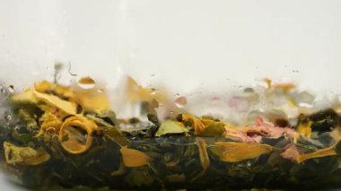 Sıcak su akıntısı şeffaf bir çaydanlığa akar. Çay yaprakları ve çiçek yapraklarıyla..