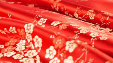 Şatafatlı kırmızı ipek kumaş arka planda çiçek desenli, doğu tarzı işlemeli, erik çiçeği desenli. İpek kumaşların kıvrımları dönen bir vitrinde güzel bir şekilde parıldıyor..