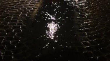Gece yağmur yağarken su birikintisindeki bir sokak lambasından yansıyan ışık. Şehirde bir kaldırım taşı caddesi.