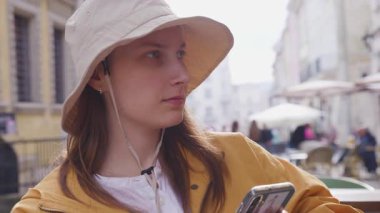 Şapkalı genç bir kız bir sokak kafesinde oturuyor, akıllı telefonu elinde tutuyor ve etrafına bakıyor..