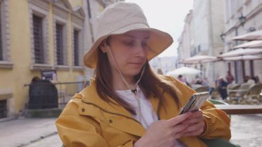 Şapkalı genç bir kız bir sokak kafesinde oturuyor ve akıllı telefonuna mesajlar yazıyor..