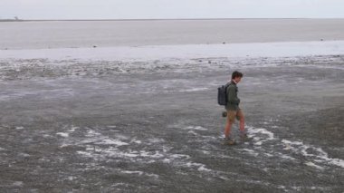 MONT-SAINT-MICHEL, NORMANDY, FRANCE - 15 Nisan 2025: İngiliz Kanalı 'nda alçak gelgit sırasında elinde kamerayla kıyı boyunca yürüyen genç bir adam.
