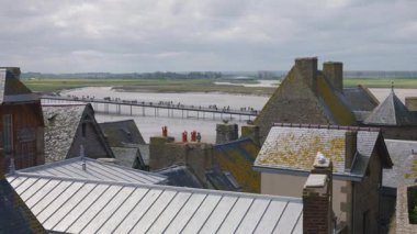 Mont-Saint-Michel Manastırı 'nın duvarlarından eski binaların kiremitli çatılarının, Fransız bayrağının, yeni köprünün ve turistlerin ve rüzgarlı bir bahar gününde Manş Denizi' nin aşağısında.