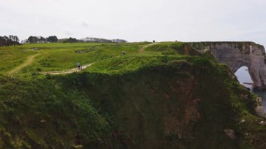 Panoramik beyaz tebeşir kayalıkları manzarası, yeşil yamaçlar, doğal kemerler ve Le Manneporte kayalıkları arasında yürüyen turistler bahar güneşli bir günde Etretat Tebeşir Kompleksi, la Manche Kanalı (İngiliz Kanalı), Normandiya, Fransa.