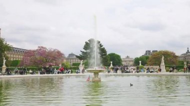PARIS, FRANCE - 21 Nisan 2025: İnsanlar 1. bölgedeki bulutlu bir bahar gününde Jardin des Tuileries 'de çiçek açan ağaçlara ve Arc de Triomphe du Carrousel' e tepeden bakan bir çeşmeyle Grand Bassin Rond 'da yürüyüp rahatlıyorlar..