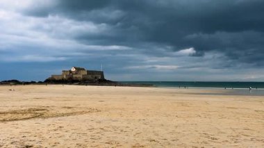 Saint Malo, Brittany, Fransa 'da fırtınalı bir gökyüzü altında güneşle aydınlatılan kumlu sahil, İngiliz Kanalı ve Ulusal Kale manzarası.
