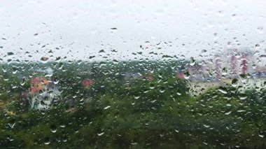 Yağmur sırasında pencere camlarından su damlaları düşüyor. Orman ağaçlarının ve şehir binalarının arka planına karşı..