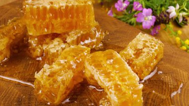 Altın bal akıntıları damlıyor ve bal peteğinin üzerine dökülüyor. Tahta bir meşe ağacından kesilmiş tahtanın üzerinde vahşi yaz çiçeklerinin arka planına karşı. Balmumu bal peteklerine yakın çekim. Sağlıklı yeme kavramı.