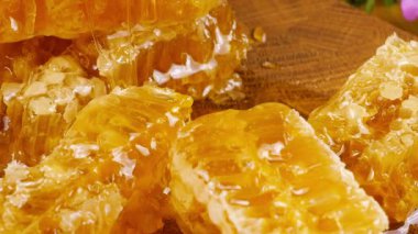 Altın bal akıntıları damlıyor ve bal peteğinin üzerine dökülüyor. Tahta bir meşe ağacından kesilmiş tahtanın üzerinde vahşi yaz çiçeklerinin arka planına karşı. Balmumu bal peteklerine yakın çekim. Sağlıklı yeme kavramı.