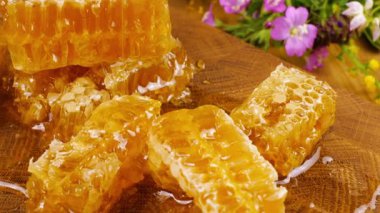 Altın bal akıntıları damlıyor ve bal peteğinin üzerine dökülüyor. Tahta bir meşe ağacından kesilmiş tahtanın üzerinde vahşi yaz çiçeklerinin arka planına karşı. Balmumu bal peteklerine yakın çekim. Sağlıklı yeme kavramı.