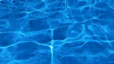 Güneşli bir günde, açık mavi suyun yüzeyinde karo kaplı ve ışık dalgalanmaları olan havuzun alt kısmının üst görüntüsü.