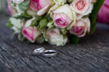 Beyaz altın alyanslar, arka planda düğün buketi olan eski bir tahta tabakta.